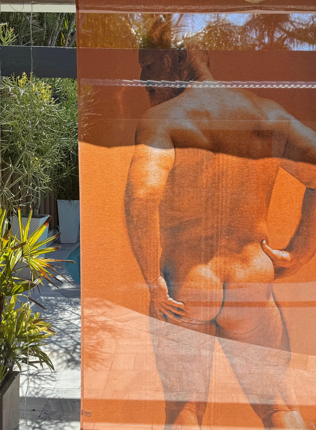 An orange mesh privacy screen with a large photographic print of a muscular man, displayed on a patio next to a swimming pool.